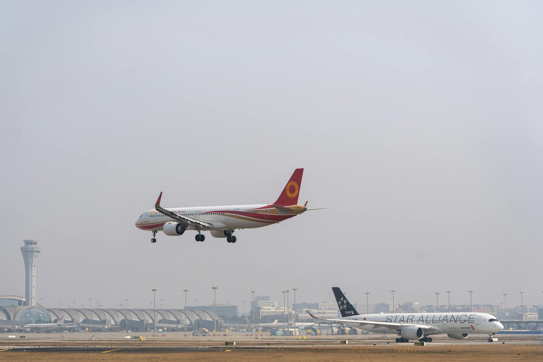 Two commercial airplanes approaching landing at a busy international airport with control tower visible.