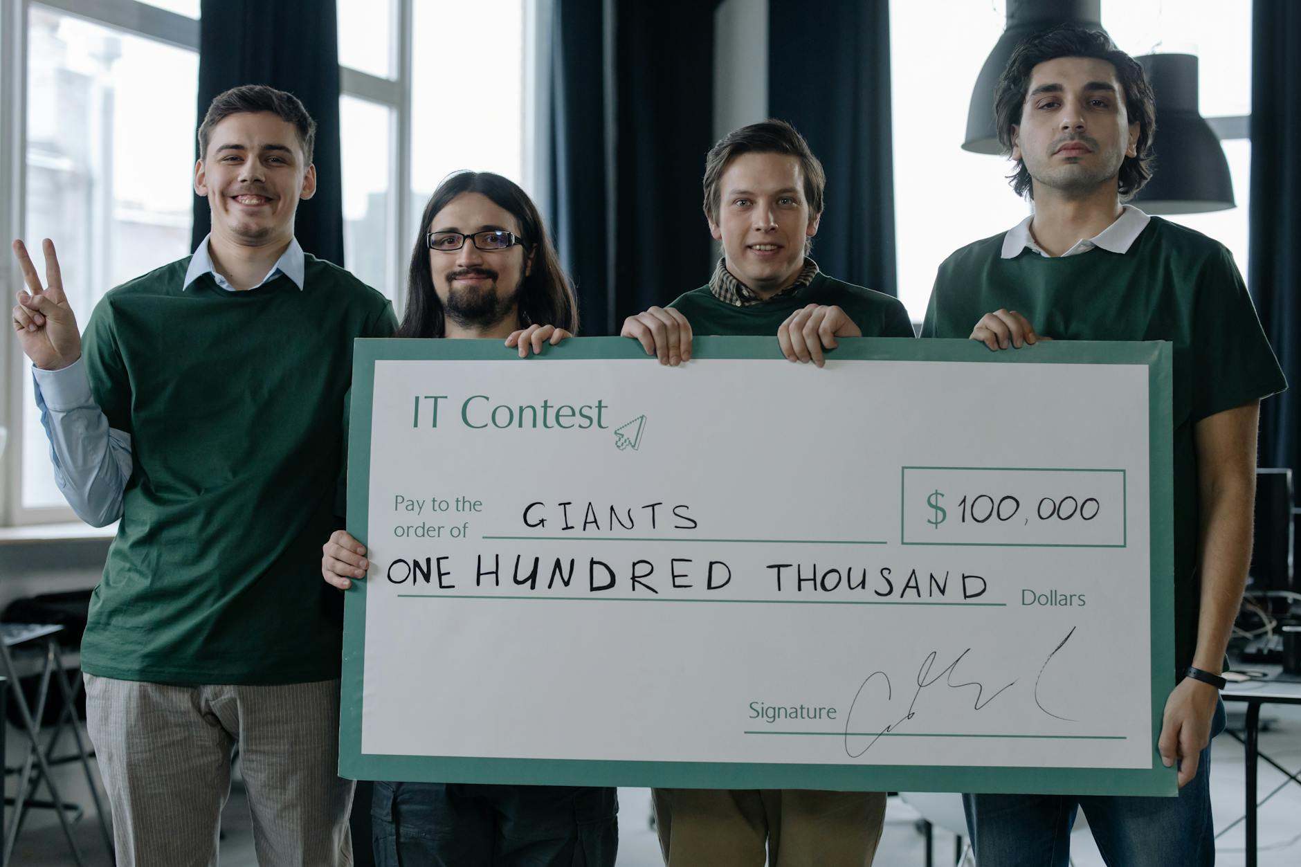 Four men celebrate a $100,000 IT contest victory holding a large cheque inside a modern office.